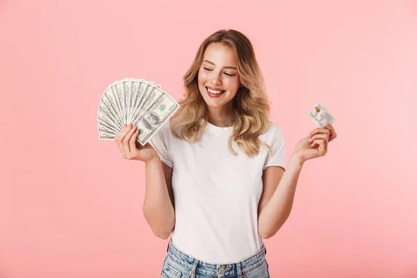 Emocionada joven rubia posando aislada sobre fondo de pared rosa sosteniendo dinero y tarjeta de crédito . — Foto de Stock