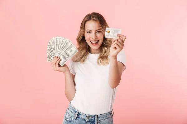 Emocionada joven rubia posando aislada sobre fondo de pared rosa sosteniendo dinero y tarjeta de crédito . — Foto de Stock