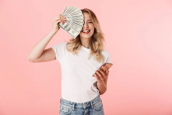Jovem loira excitada posando isolada sobre fundo de parede rosa segurando dinheiro e usando telefone celular . — Fotografia de Stock