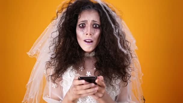 在万圣节穿着婚纱和化妆使用智能手机和看着橙色背景的相机 — 图库视频影像
