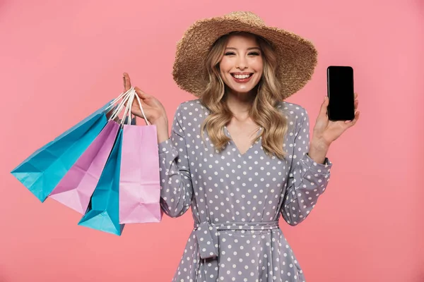 Increíble joven posando aislada sobre fondo de pared rosa sosteniendo bolsas de compras y mostrando la pantalla del teléfono móvil . —  Fotos de Stock