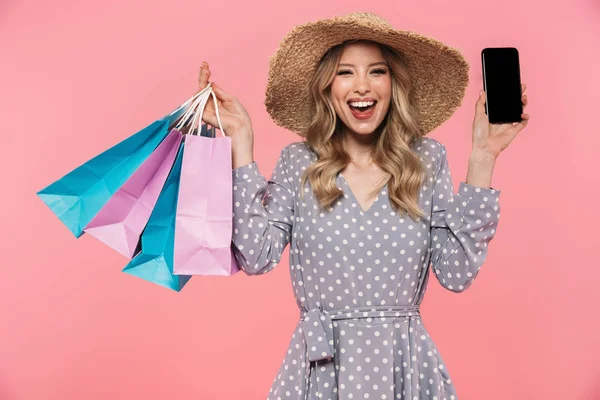 Incrível jovem posando isolado sobre fundo de parede rosa segurando sacos de compras e mostrando exibição de telefone celular . — Fotografia de Stock