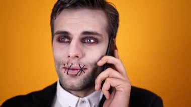 Turuncu arka plan üzerinde akıllı telefon ile konuşmaya çalışırken dikişli ağız ile cadılar bayramı makyaj karışık ölü damat