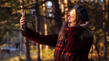 Parkta dururken akıllı telefondan selfie çeken bere şapkalı ve paltolu mutlu esmer kadın
