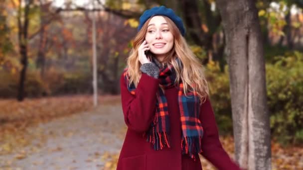 公園を歩きながらスマホで話すベレー帽とコートを着た陽気なブルネットの女性 — ストック動画