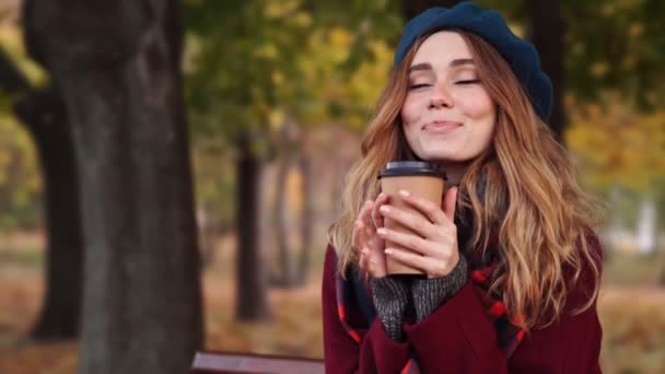 公園のベンチに座りながら ベレー帽をかぶった笑顔のブルネット女性のクローズアップビューとコーヒーを飲むコート — ストック動画