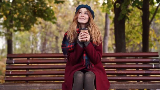 公園のベンチに座っている間 ベレー帽とコートでコーヒーを飲んで笑顔のブルネットの女性 — ストック動画