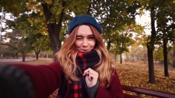 公園のベンチに座っている間に自分撮りを作るベレー帽とコートで幸せなブルネットの女性 — ストック動画