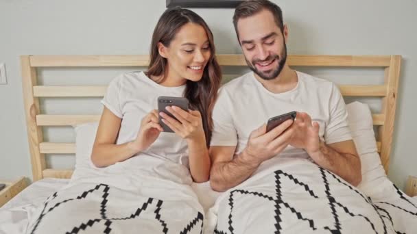 Ευτυχής Υπέροχο Ζευγάρι Χρησιμοποιώντας Smartphones Ενώ Ξαπλωμένοι Μαζί Στο Κρεβάτι — Αρχείο Βίντεο