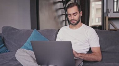 Evde kanepede otururken dizüstü bilgisayar kullanarak Ciddi yakışıklı sakallı adam