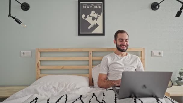 寝室でベッドに横たわっている間 ラップトップコンピュータを使用して陽気なあごひげの男 — ストック動画
