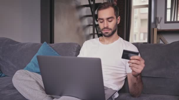 気楽なハンサムなひげの男は 自宅でソファに座っている間 ラップトップコンピュータ上の請求書を支払います — ストック動画