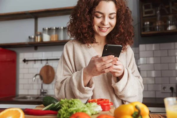 Foto de una mujer caucásica feliz sosteniendo el teléfono inteligente mientras cocina — Foto de Stock