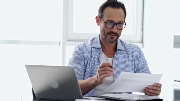 穿着衬衫和眼镜的熟男人坐在桌子旁 在家里用笔记本电脑和文件 — 图库视频影像