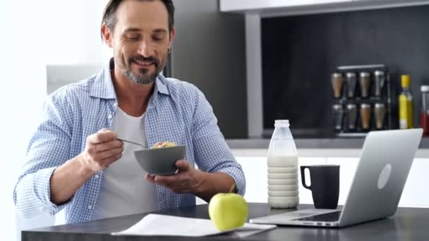 自宅で朝食を食べながら ラップトップコンピュータでテーブルのそばに座っているシャツの幸せな成熟した男 — ストック動画