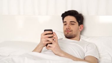 Yakışıklı esmer smiing adam yatakta yatan ve akıllı telefon kullanarak rahat giysiler giyiyor