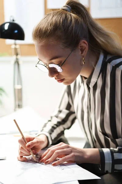 Fotó a fókuszált európai nő építész visel szemüveg tervezése tervezetet, és ül a munkahelyen — Stock Fotó