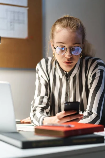 Foto de una mujer arquitecta caucásica que usa anteojos usando un teléfono celular y está sentada en el lugar de trabajo — Foto de Stock