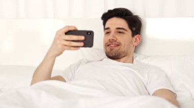 Yatakta yatan ve akıllı telefonda selfie yapma rahat giysiler içinde Yakışıklı esmer neşeli adam 