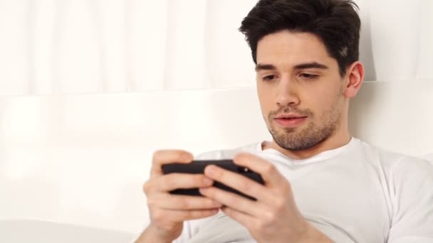 集中的黑发打人穿休闲衣服玩智能手机游戏 并赢得 而躺在床上 — 图库视频影像