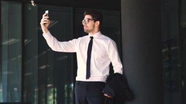 Cam binanın yanında dururken akıllı telefondan selfie çeken resmi takım elbise ve gözlük giymiş mutlu işadamı
