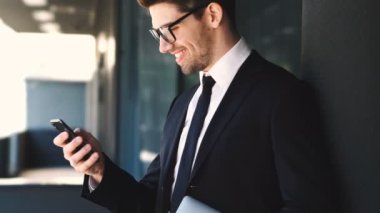 Cam binanın yanında dururken akıllı telefon kullanarak resmi takım elbise ve gözlük giymiş gülümseyen işadamı