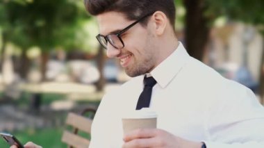 Parkta bankta otururken akıllı telefon ve kahve içerken resmi takım elbise ve gözlük giymiş Mutlu işadamıyan görünümü
