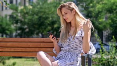 Parkta bankta otururken akıllı telefon kullanarak elbise Pretty memnun sarışın kadın
