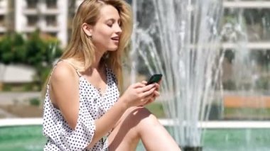 Parkta çeşme yakınında otururken akıllı telefon kullanarak elbise memnun sarışın kadının yan görünümü