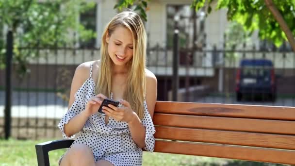 快乐的金发女人在礼服使用智能手机 而坐在公园的长凳上 — 图库视频影像