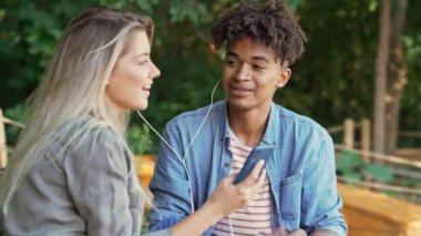 Mutlu genç çok ırklı çift parkta birlikte vakit geçirirken akıllı telefon ve şarkı ile müzik dinlerken