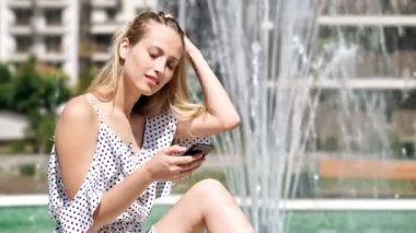 Parkta çeşme yakınında otururken akıllı telefon kullanarak elbise oyuncu sarışın kadın Yan görünümü