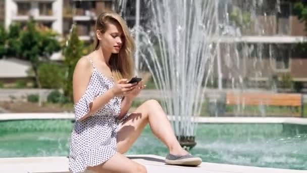 Радісна Блондинка Одязі Використовує Смартфон Сидячи Біля Фонтану Парку — стокове відео