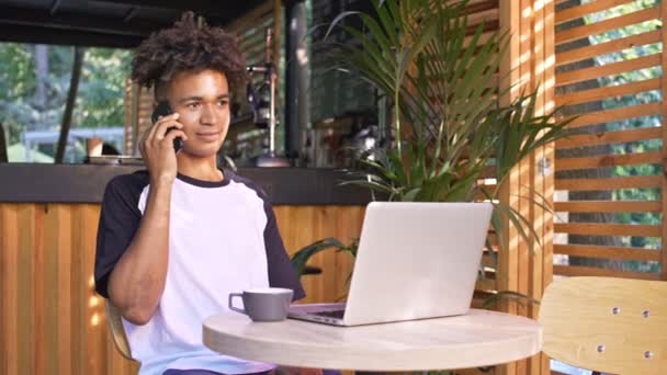 Улыбающийся Африканский Студент Разговаривает Смартфону Сидя Столом Ноутбуком Кафе Открытом — стоковое видео