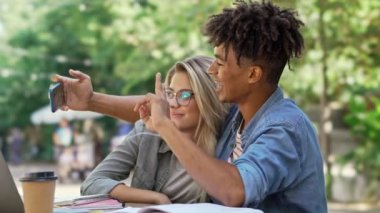 Neşeli genç çok ırklı çift şehir kafede dizüstü bilgisayar ile masanın yanında otururken akıllı telefonda selfie yapıyor