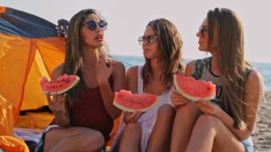 Bir grup mutlu arkadaş karpuz yiyor ve denize yakın plajda birlikte eğleniyor