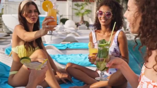 Gruppe fröhlicher Freundinnen in Badeanzügen, die Cocktails trinken, während sie es sich auf Liegestühlen in der Nähe des Swimmingpools gemütlich machen