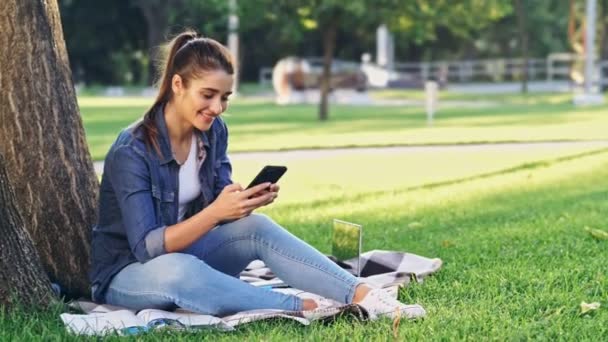 Mujer Morena Sonriente Usando Teléfono Inteligente Mientras Está Sentado Cerca — Vídeo de stock