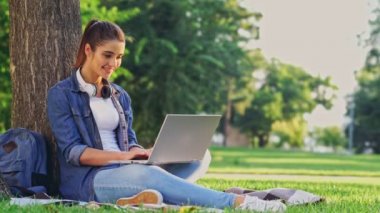 Parkta çim ağacın yanında otururken dizüstü bilgisayar kullanarak Mutlu esmer kadın