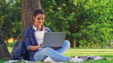 Parkta çim ağacın yanında otururken dizüstü bilgisayar kullanarak memnun esmer kadın