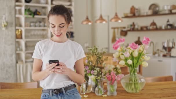 ダイニングルームのテーブルの近くに立っているスマートフォンを使用して白いTシャツを着たかなり若いブルネットの女性 — ストック動画