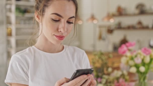 ダイニングルームに立ってスマートフォンでおしゃべり白いTシャツを着たかわいい若いブルネットの女性のクローズアップビュー — ストック動画