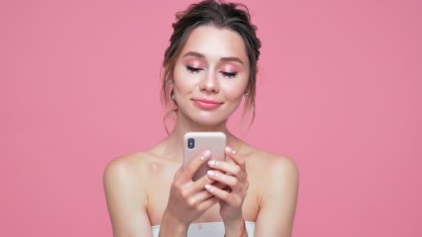 ピンクの背景の上にスマートフォンを使用して顔と手に真珠を持つ美しさ幸せなブルネットの女性 — ストック動画