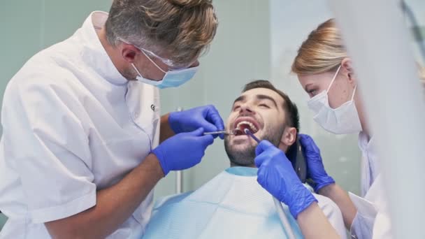 歯科医と歯科医とアシスタントと歯科治療を受けながら微笑む美しいブルネットの男 — ストック動画