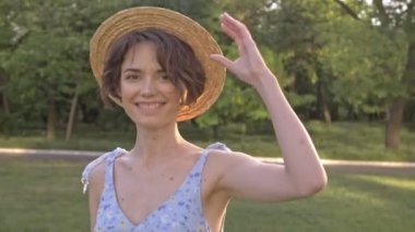 Yeşil parkta yürürken gülümseyen ve hasır şapka çıkararak çekici genç kadın