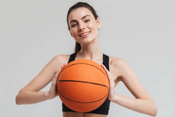 Mladá sportovní fitness žena hráč dělá cvičení basketbalovaná přes šedou zeď pozadí. — Stock fotografie