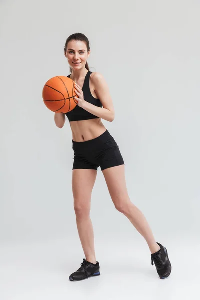 Mladá sportovní fitness žena hráč dělá cvičení basketbalovaná přes šedou zeď pozadí. — Stock fotografie