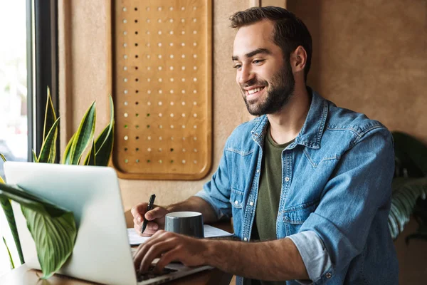 Фото усміхненого бородатого чоловіка, який пише і друкує на ноутбуці під час роботи в кафе в приміщенні — стокове фото