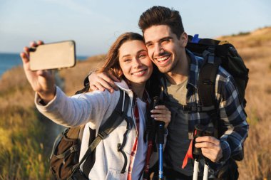 Ücretsiz alternatif tatil kamp sırt çantası ile dışarıda Mutlu genç sevgi dolu çift cep telefonu ile selfie çekmek.