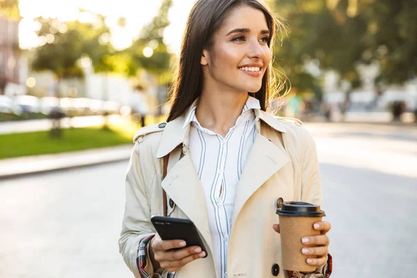 Retrato de mujer morena caucásica bebiendo café para llevar y escribiendo en el teléfono celular mientras camina por la calle de la ciudad — Foto de Stock